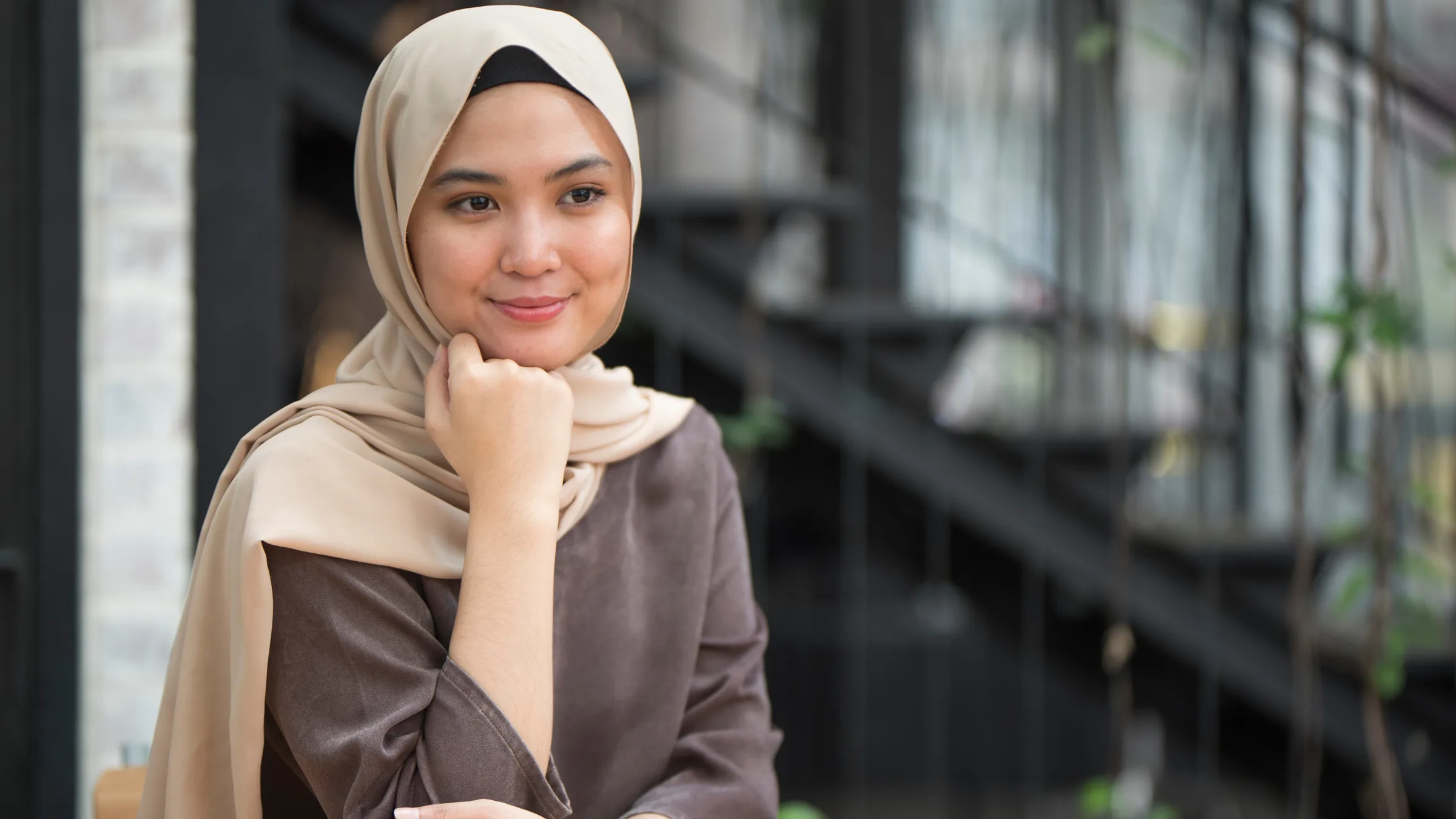 A customer in a hijab seated at a cafe, resting her chin on her hand and smiling